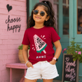 A young girl wearing round gold-framed sunglasses, a maroon graphic t-shirt with a popsicle design and the text 'BE COOL KIND', and white shorts, posing in front of a pink brick wall with 'choose happy' written on it.