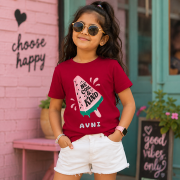 A young girl wearing round gold-framed sunglasses, a maroon graphic t-shirt with a popsicle design and the text 'BE COOL KIND', and white shorts, posing in front of a pink brick wall with 'choose happy' written on it.
